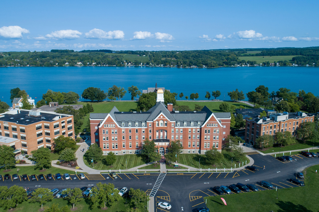 Aerial view of a large red-brick academic building on a lakeside campus, with surrounding parking lots, green lawns, and blue water stretching across the background under a partly cloudy sky.