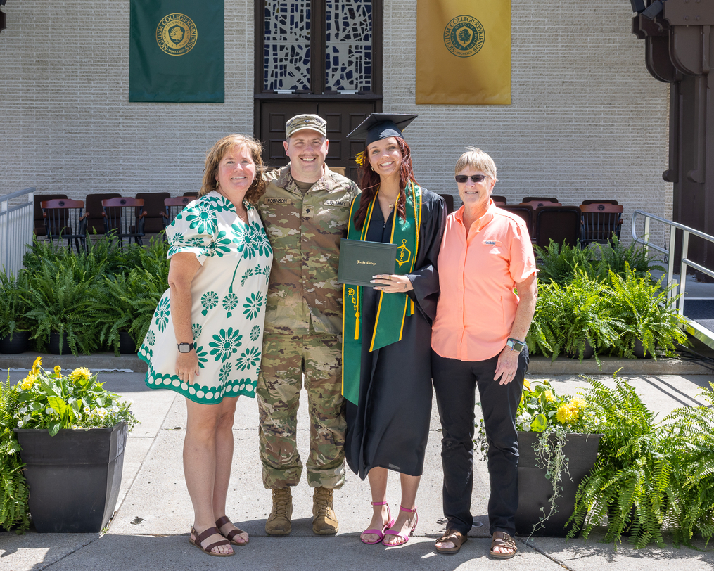 A graduate in cap and gown poses with family members and a person in military uniform while holding a diploma cover on the Keuka College Commencement stage.