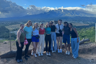 Group photo of eight people standing on a mountain overlook with dramatic clouds and a valley landscape behind them.