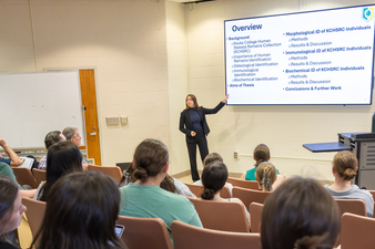 A student presenter stands at the front of a classroom, gesturing toward a large screen displaying a slide titled “Overview.” The slide outlines topics including background, methods, and conclusions related to human skeletal remains research. The presenter wears professional attire and faces an audience of seated students in a lecture hall, who are watching attentively. A whiteboard and classroom equipment are visible along the walls.